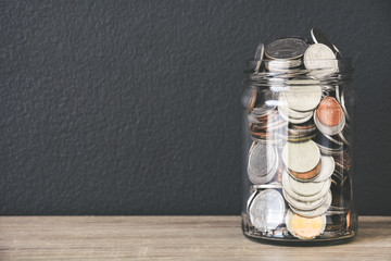 transparent glass savings jar filled with coins on wooden table with black color wall background, copy space