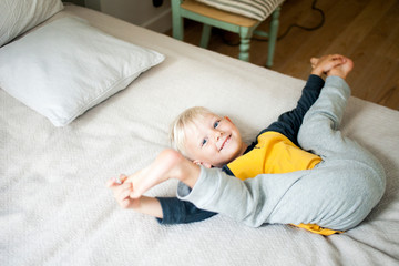 A six-year-old blond boy lies in bed holding his legs with his hands looks into the frame.