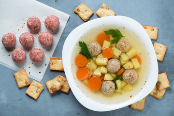 Plate of meatball soup over grey concrete background with crackers and raw meatballs, flatlay, horizontal shot