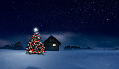 Glänzend leuchtender Christbaum im Schnee bei einer Holzhütte mit leuchtendem Fenster