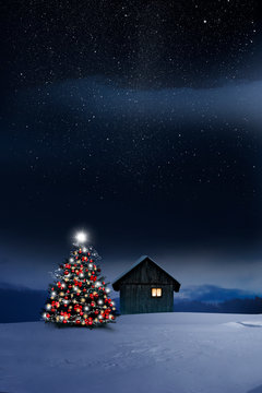 Gl&auml;nzend leuchtender Christbaum im Schnee bei einer Holzh&uuml;tte mit leuchtendem Fenster