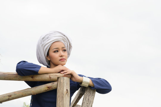 Beautiful Young Asian Woman Wearing Traditional Dress With Turban Sitting On Wooden Terrace With Sad Face Looking Up Thinking Over White Cloud Sky Background.