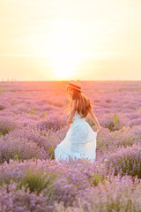 Fototapeta na wymiar Beautiful slim girl in white dress and hat whirls in lavender field at sunset in summer