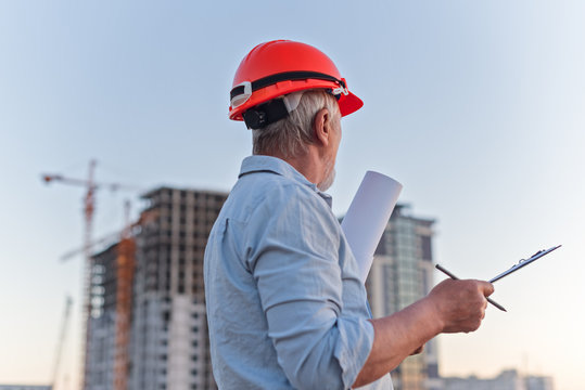 Constructor engineer with a gray beard in a constractor helmet with drawings on the background of construction