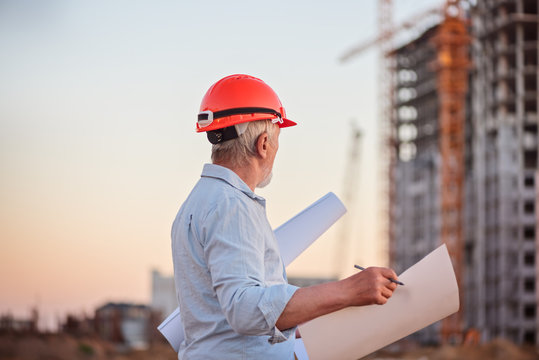 Constructor engineer with a gray beard in a constractor helmet with drawings on the background of construction