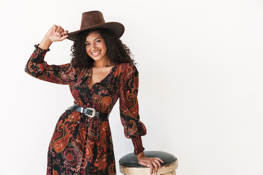 Image of young african american cowgirl in dress and hat standing by chair