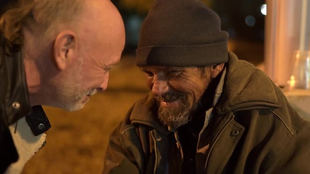 Passerby talking to homeless man on the sidewalk at night.