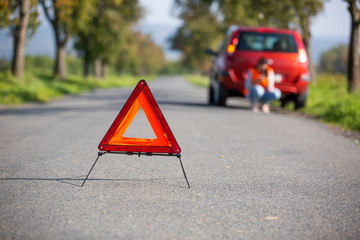 Annoyed young woman in a road distress situation - setting up a warning triangle and calling for assistance after her car broke down in the middle of nowhere; transportation concept (car problem)