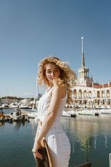 A swift wind blowed hair over the face of a pretty blonde who stands stretched out like a string and turned her head to shoulder against the background of the seaport and water