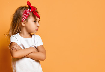 Kid girl in white t-shirt and red headband is standing with her arms crossed, looking aside at copy space on yellow 