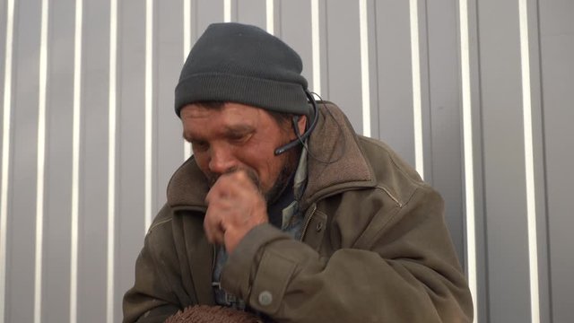 Sad homeless man listening to music with earphones and mobile phone. Wretched beggar singing along with the song on smart phone.