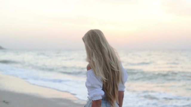 Back view of beautiful slim blonde with long hair in a white blouse walkng along the coast at sunset