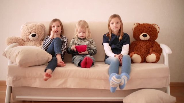 Funny little boy (3 years old) watches cartoons with older sisters (8 and 9 years old). Brother and sisters are sitting on the couch and eating cookies.