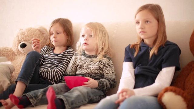 Funny little boy (3 years old) watches cartoons with older sisters (8 and 9 years old). Brother and sisters are sitting on the couch and eating cookies.