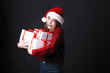 Lovely girl with make-up in a burgundy sweater and santa claus hat. Posing on a dark background holds gifts on a Christmas tree, the concept of Christmas and New Year