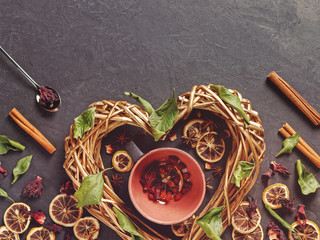 Romantic composition for Valentine's Day. A wicker heart is on a dark background. Inside it is an empty mug with herbs and berries for making a healthy drink. Nearby are various dried herbs and plants