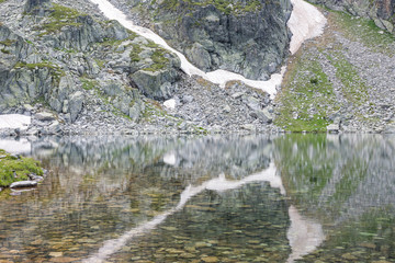 Lake stone shore reflected in calm water. Last snow left in summer in the mountain. Symmetry. Selective focus. Texture, background.