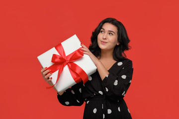 Brunette woman in a black dress smiling holding Christmas giftbox of white color with red ribbon.