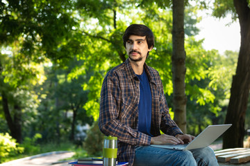 Young brunet freelancer with beard and mustache sitting with a laptop and coffee in a park.