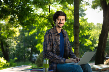 Fototapeta premium Young brunet freelancer with beard and mustache sitting with a laptop and coffee in a park.