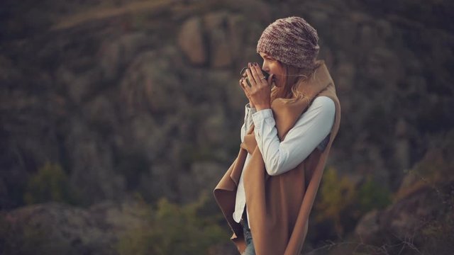 Happy beautiful woman with blond hair in a hat and waistcoat drinks hot tea from an iron cup, on the background of a canyon, the concept of outdoor activities and travel