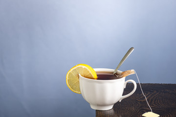 white mug with tea and bag, lemon and a spoon inside on the edge of the table on a gray background