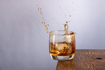 ice falls into a glass standing on the edge with whiskey and splashing on a gray background