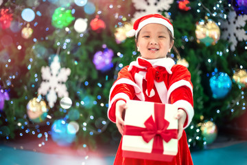 Cute little girl in red Santa Claus outfit holding a Christmas present in her hands on the background of Christmas tree.