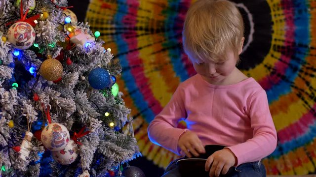Cute blond boy in pink sweatshirt holds case for glasses in his hands sitting near Christmas tree, colored background. child takes glasses out of case, puts them on and packs them back. Shallow focus