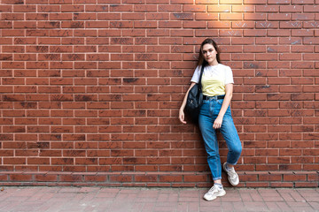 Fototapeta na wymiar Attractive serious young brunette girl in blue jeans and yellow t-shirt, with black shoulder bag isolated over red brick wall