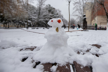 Little funny snowman on a bench in a city park. snow woman with red eyes. New Year's holidays. winter fun