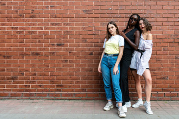 Full length stylish portrait of the young beautiful multi ethnic girls in casual clothes isolated over red brick wall