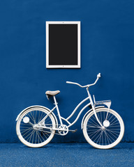 A white color painted bicycle stands against a blue wall. A white frame with a black empty space for inscriptions and pictures hangs on the wall. Minimalistic classic concept for creative ideas.