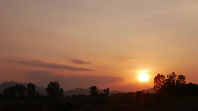 Time lapse of evening golden sky cloudscape turn red-orange shade sunset, the sun goes down below the horizon behide mountains and silhouette forest trees in countryside landscape.