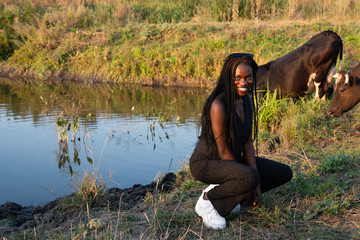 Happy smiling african girl in black clothes crouched and looking at the camera, cow drinks water...