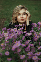  Portrait of a young woman with curly white hair in a black sweater and in a wreath on the street in a summer park on a background of green trees. Purple flowers in the foreground