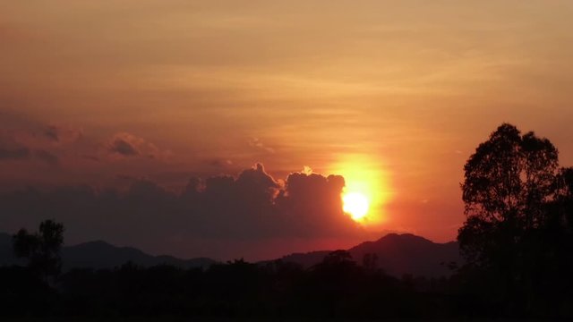 Time lapse of evening golden sky cloudscape turn red-orange shade sunset, the sun goes down below the horizon behide mountains and silhouette forest trees in countryside landscape.