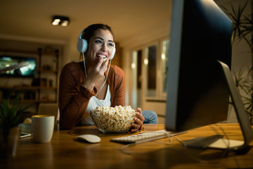 Happy woman eating popcorn and watching a movie on desktop PC at night. - obrazy, fototapety, plakaty