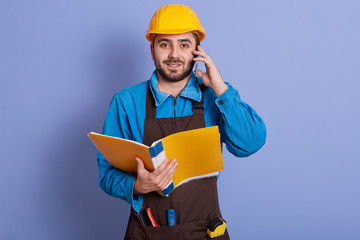 Close up portrait of young attractive bearded builder having conversation via phone and holding paper folder in hands, dresses blue shirt, brown apron, yellow helmet. Buildingand engineering concept.
