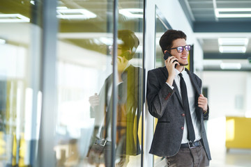 Young businessman in glasses talking on cell phone standing in the modern office. Stylish man in a suit with folder standing and talking on telephone. Communication concept.