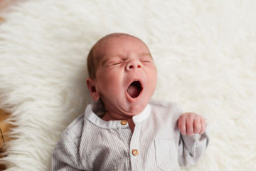 bebe de 3 semanas (menos de un mes) bostezando, bostezo de niño pequeño tumbado en alfombra de pelos blancos y vestido de lino.