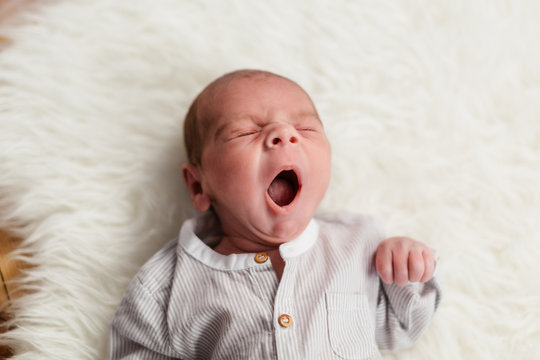 bebe de 3 semanas (menos de un mes) bostezando, bostezo de ni&ntilde;o peque&ntilde;o tumbado en alfombra de pelos blancos y vestido de lino.