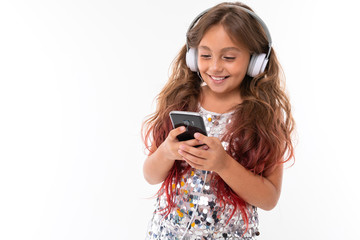 Little pretty caucasian girl listen to music with big earphones and serfing internet with phone, picture isolated on white background