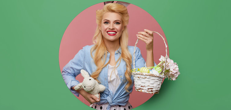 Young pin-up woman with easter bunny ears with a basket of colored eggs crawls out through a round hole in the wall. Menthol or turquoise color background. Holiday card blank