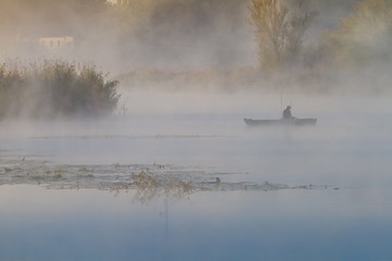 unknown fisherman in a punt boat fishing with rods, thick fog covers still surface of a river, bulrush, cane and willows grow on a bank, water steam on a cold autumn morning, generous nature