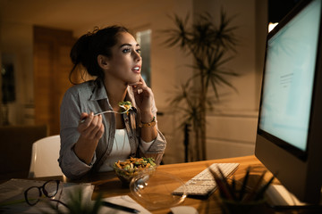 Young woman reading an e-mail on desktop PC while eating salad at home.