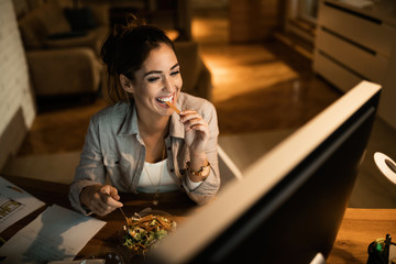 Happy woman eating salad while working on desktop PC at night.
