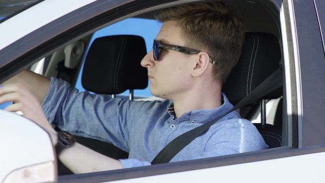 Brunette man in casual clothes and sunglasses fastens his seat belt and driving car with bluesceen background. He is sitting in his white car with the greenscreen background