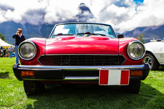 Classic Old American Red Car Front Low Angle View, Headlights Light Lamps And A Black Grille With Chrome Bumper, During Outdoor Antique Cars Show