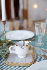 Cup for hot holidays beverage on the table served for the Christmas brunch with beautiful festive dishes, candelabra with candles and fir branches. Christmas eve.